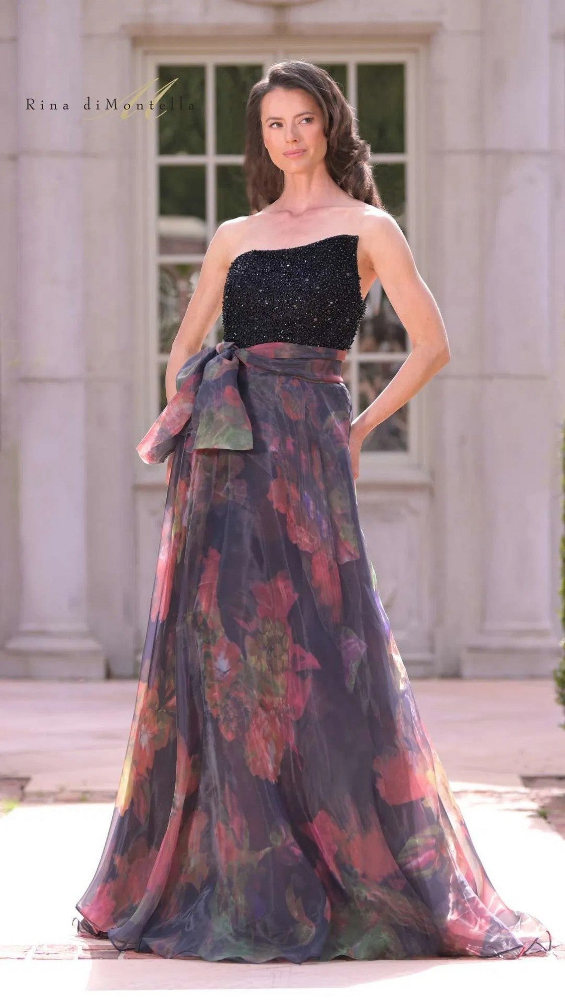 A woman stands outdoors in front of a building wearing the Long Formal Dress: Rina Di Montella RD3213—a sleeveless black and multicolored floral gown with an organza skirt, sheer overlay, and large bow.